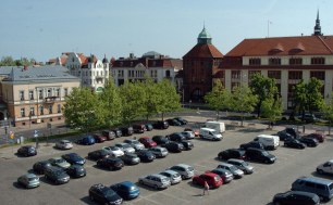 na zdjęciu widzimy plac, kilkadziesiąt samochodów zaparkowanych, kilka drzew, w tle jednię i inne budyni